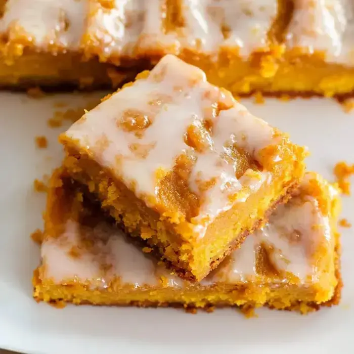 A close-up of a moist, glazed pumpkin dessert cut into squares, with a few pieces arranged on a white plate.