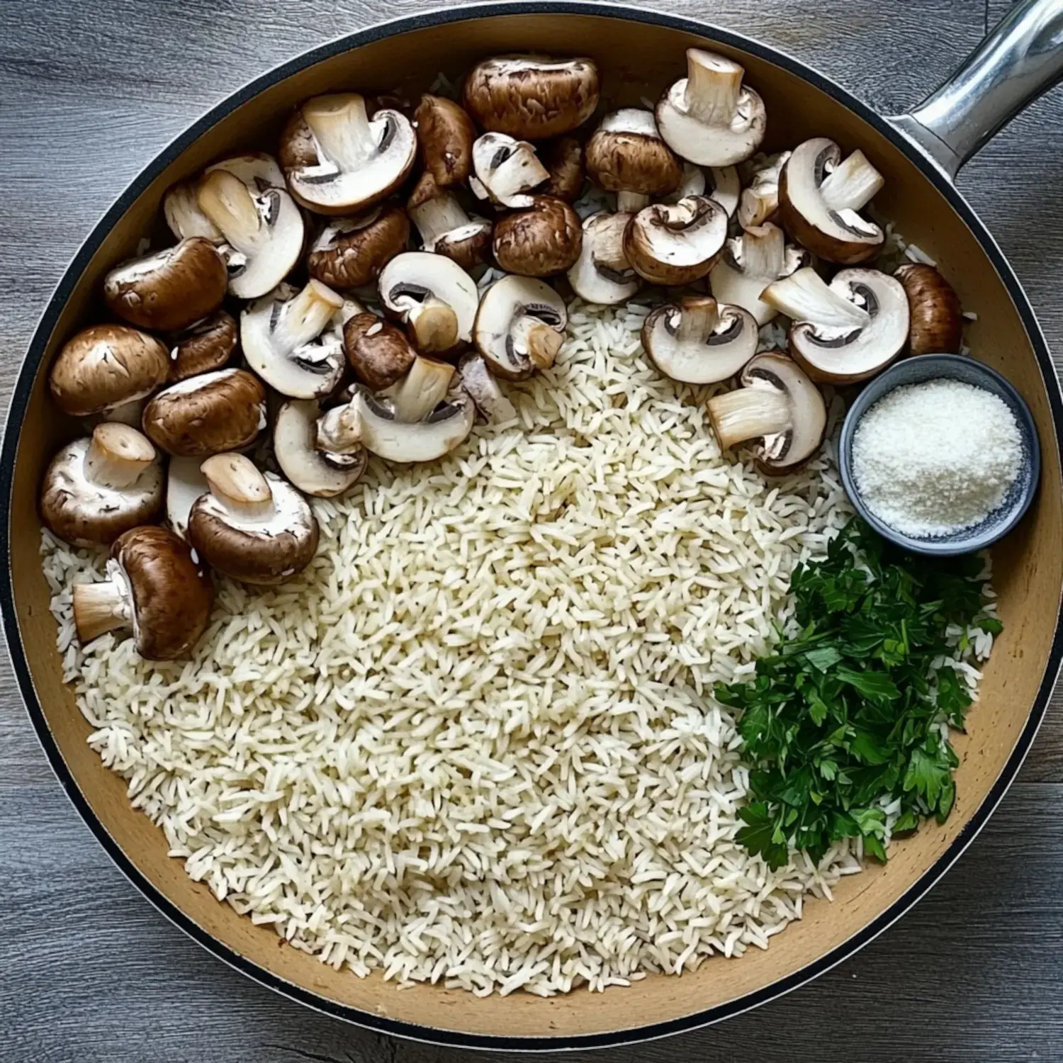 A skillet contains a mixture of white rice, sliced mushrooms, chopped parsley, and a small bowl of grated cheese.