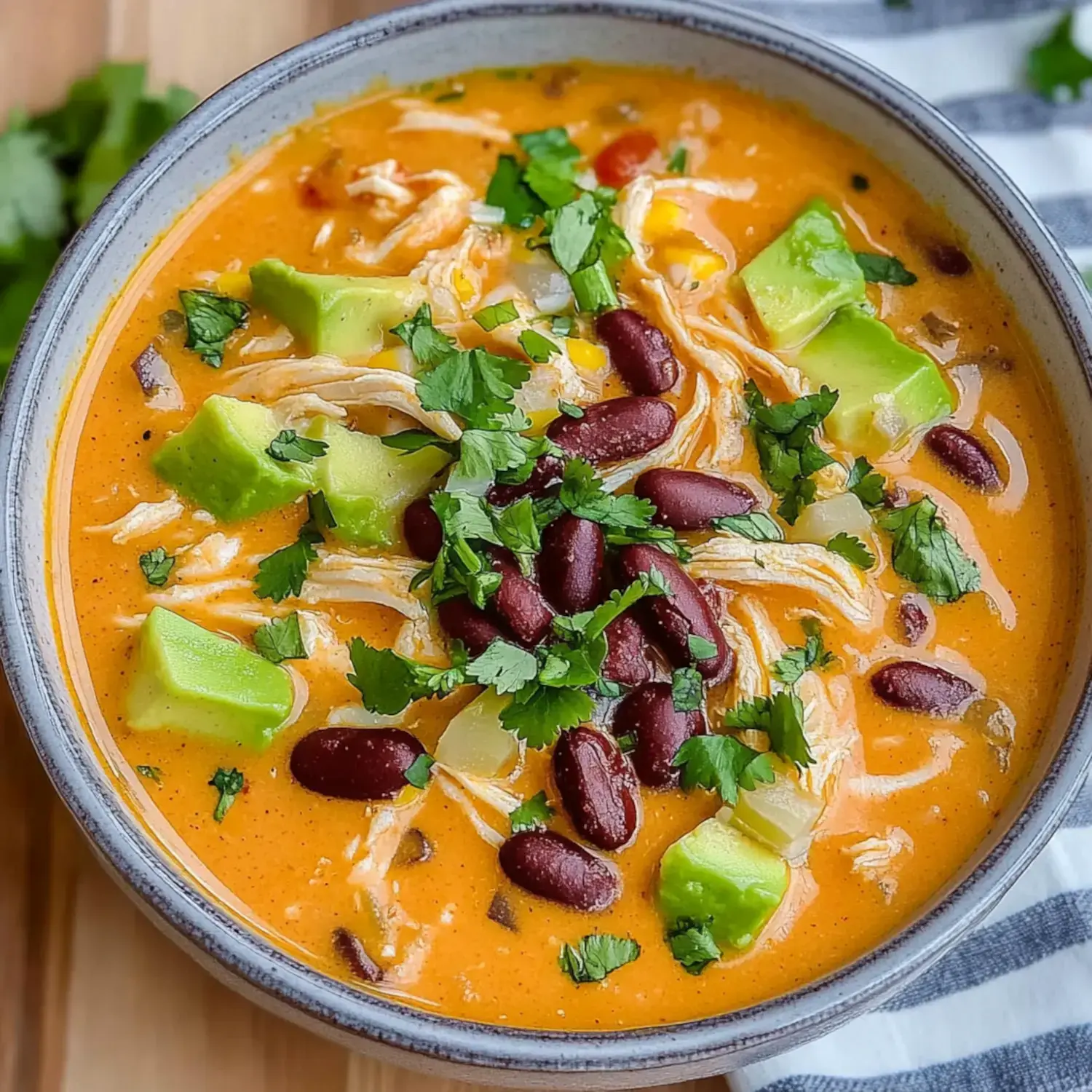 A bowl of velvety orange soup garnished with pulled chicken, chunks of avocado, red beans, yellow corn, and green cilantro sprigs.